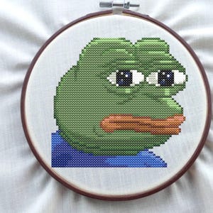 May include: Cross-stitch embroidery of a green frog face with large eyes and a brown mouth, set in a wooden embroidery hoop. The frog is stitched on white fabric, with a blue section at the bottom.