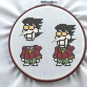 May include: A cross-stitch embroidery hoop featuring pixelated characters. The characters have black hair, yellow glasses, and are wearing red and green outfits. The embroidery is set against a white fabric background, framed by a wooden hoop.