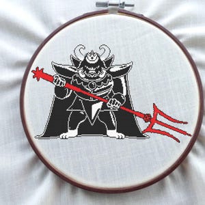 May include: A cross-stitch embroidery featuring a black and white pixelated character with a red trident. The character has horns and is wearing a cape. The embroidery is set in a wooden hoop.