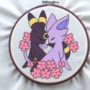 May include: Cross-stitch embroidery in a wooden hoop featuring two stylized cat-like creatures, one black and one lavender, surrounded by pink flowers. The creatures are facing each other. The embroidery is on a white fabric background.