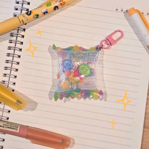 May include: A clear plastic candy bag charm with a pink clasp, filled with colorful candies and character illustrations. The bag has a rainbow-colored bottom and is resting on a lined notebook with pens.