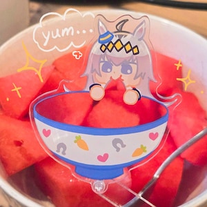 May include: A clear acrylic charm featuring a cartoon character with a bowl of watermelon. The bowl has a blue rim and is decorated with hearts, carrots, and horseshoes. The text "yum..." is in a speech bubble above the character.