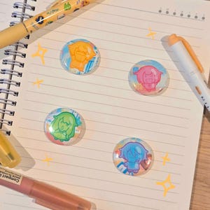 Puede incluir: Cuatro botones redondos coloridos con diseños de personajes de estilo anime. Los botones presentan personajes naranjas, rosas, verdes y azules. Los botones están sobre una página de cuaderno rayada con bolígrafos y marcadores en el fondo. Hay estrellas decorativas dibujadas en la página.
