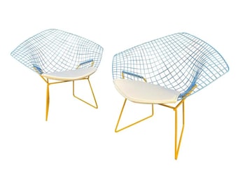 May include: Two modern diamond-shaped wire chairs. The chairs have a light blue wire frame, yellow legs, and a white cushion. The chairs are designed with a unique, open-air aesthetic, perfect for indoor or outdoor use.