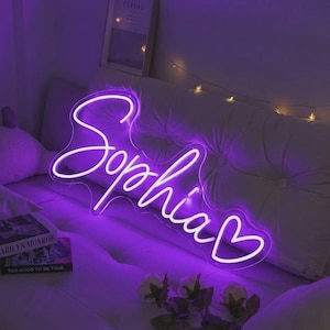 Peut inclure: Enseigne au néon blanche en forme du prénom "Sophia" écrit en cursive, avec un cœur à la fin, sur fond violet. L'enseigne est posée sur une surface blanche. L'ensemble dégage une ambiance douce et lumineuse.