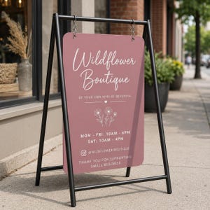 May include: A black metal A-frame sidewalk sign with a dusty rose rectangular board. The sign features white text reading Wildflower Boutique, Be Your Own Kind Of Beautiful, business hours, an Instagram handle, and a line drawing of three daisies.