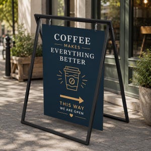 May include: A black metal A-frame sidewalk sign with a dark blue rectangular board. The board features white and gold text reading COFFEE MAKES EVERYTHING BETTER, a gold coffee cup icon, a gold arrow, and the text THIS WAY WE ARE OPEN.