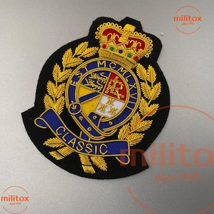 以下が含まれることがあります： 金色の王冠、月桂樹の葉、中央のエンブレムが付いた黒色の刺繍パッチ。エンブレムは、青いバナーに「CLASSIC」という言葉が入った青と黄色のデザインが特徴です。パッチには「EST MCMLXVII」というテキストがあります。