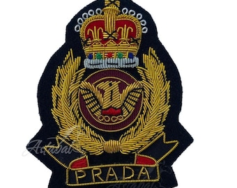 May include: Embroidered patch featuring a gold crown, laurel leaves, and a central emblem on a black background. The word "PRADA" is embroidered in gold at the bottom. The patch has a detailed, textured appearance.
