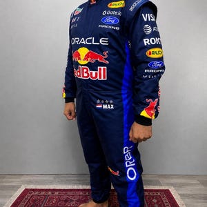 May include: A dark blue racing suit featuring the Red Bull logo, Oracle branding, and Ford Racing text. The suit includes various sponsor logos, four stars, a Dutch flag, and the name MAX printed on the chest, with blue side panels and black cuffs.