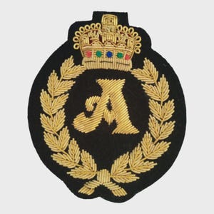 May include: Black embroidered patch featuring a gold letter "A" within a gold laurel wreath. A gold crown with red, green, and blue accents sits above the letter. The patch has a black background and is likely a decorative emblem.