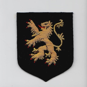 May include: Embroidered patch featuring a gold lion rampant with red claws and tongue on a black shield-shaped background. The lion is highly detailed, with textured gold thread. The patch has a classic heraldic design.