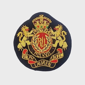 Könnte beinhalten: Ein kreisförmiger, dunkelblauer Aufnäher mit Goldstickerei, der eine Krone über zwei aufsteigenden Löwen zeigt, die einen roten Mittelkreis mit den Buchstaben RL flankieren. Das Design enthält ein Banner mit dem Text ESTABLISHED MCLXVII RALPH LAUREN.