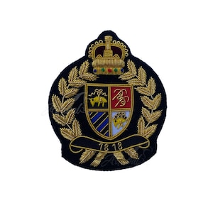 Op de afbeelding: Een marineblauw geborduurd wapenschild met een gouden kroon, laurierbladeren en een schild met een wapen. Het schild heeft een rood-goud ontwerp, met de cijfers "78 18" op een banner eronder.