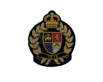 May include: A navy blue embroidered crest with a gold crown, laurel leaves, and a shield featuring a coat of arms. The shield has a red and gold design, with the numbers "78 18" on a banner below.