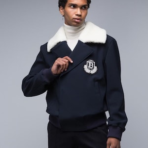 May include: A navy blue double-breasted jacket featuring a white faux-fur collar, ribbed cuffs, and a white embroidered crest patch on the chest with a letter B surrounded by laurel leaves, worn over a cream-coloured mock neck sweater.