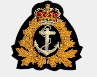 May include: A black embroidered patch featuring a gold crown, a white anchor within an oval, and gold maple leaves. The patch is likely a military or nautical insignia, with intricate detailing and a formal design.