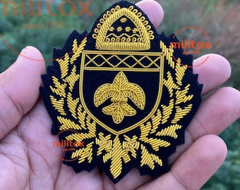 May include: A navy blue embroidered patch with gold thread detailing. The patch features a shield design with a fleur-de-lis and a crown, surrounded by laurel leaves. The text "militox since 1999" is visible.