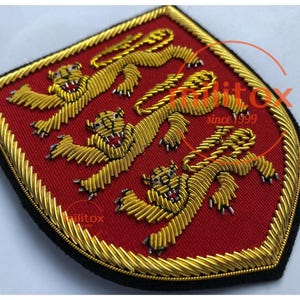 May include: Embroidered patch featuring three gold lions on a red background, framed by a gold border. The patch has a black edge and a shield shape. The lions are detailed with open mouths and outstretched paws.