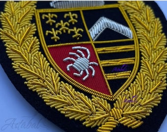 May include: Embroidered crest with a black background, featuring a shield design with gold, silver, red, and black details. The crest is surrounded by a gold laurel wreath. The shield has a crab, a silver chevron, and other symbols.