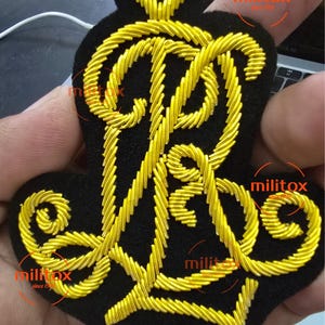 Op de afbeelding: Zwart geborduurd embleem met een goudkleurig monogram en decoratieve rand. Het monogram is de letter "R". Het embleem wordt in een hand gehouden. De tekst "militox since 1999" is zichtbaar in de afbeelding.