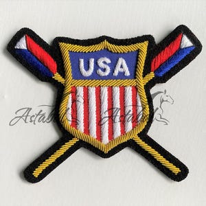 May include: Embroidered patch featuring a shield design with the text "USA" in white on a blue background. The shield has red and white stripes and is framed by gold trim and crossed black and gold bars.
