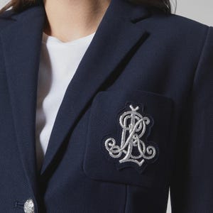 May include: A navy blue blazer with a silver embroidered emblem on the pocket. The blazer has a classic design with a notched lapel and a single button closure. The emblem features ornate script lettering.