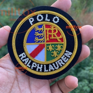 以下が含まれることがあります： 白と金色の糸で「POLO RALPH LAUREN」の文字が刺繍された円形のパッチ。パッチは、青、赤、白、緑のデザインと金の縁取りが施されたマルチカラーのエンブレムが特徴です。