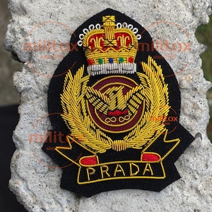 Puede incluir: Parche bordado negro con una corona dorada, un emblema de águila dorada y la palabra "PRADA" en letras doradas. El parche tiene una combinación de colores rojo y dorado y está sobre un fondo gris texturizado.
