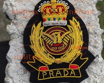 May include: A black embroidered patch featuring a gold crown, a gold eagle emblem, and the word "PRADA" in gold lettering. The patch has a red and gold color scheme and is set against a textured gray background.