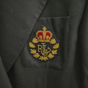 May include: Close-up of a black blazer with a decorative patch on the pocket. The patch features a gold crown with red and green accents above the letters "RL" in gold. The blazer's fabric appears to be a solid, dark color.