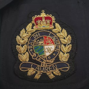 May include: A black fabric patch featuring an embroidered Ralph Lauren crest. The design includes a gold crown with red accents, a central shield divided into four quadrants with red, white, blue, and green colors, and gold laurel leaves.