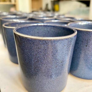 Könnte beinhalten: Eine Sammlung handgefertigter, zylindrischer Keramikbecher. Die Becher sind tiefblau mit einer gesprenkelten Textur und einem helleren Rand. Die Becher sind in Reihen angeordnet.