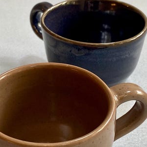 Peut inclure: Deux tasses en céramique avec anses. L'une est bleu marine profond avec un bord doré, l'autre est marron clair. Ces tasses sont pour le café ou le thé.