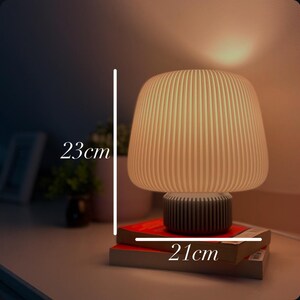 May include: A beige mushroom-shaped table lamp, 23 cm tall and 21 cm wide, with a ribbed lampshade and a dark grey base. The lamp is placed on top of two books with red covers.
