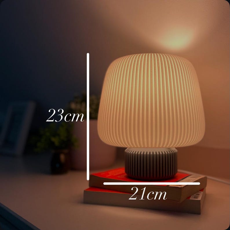 May include: A beige mushroom-shaped table lamp, 23 cm tall and 21 cm wide, with a ribbed lampshade and a dark grey base. The lamp is placed on top of two books with red covers.