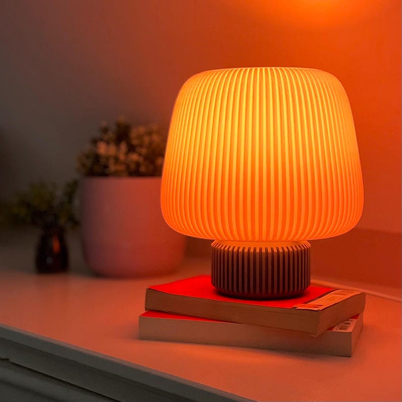 May include: A warm-toned table lamp with a ribbed, mushroom-shaped shade. The lamp sits on a dark brown base, resting on two books with red covers. The background features a soft, orange glow.