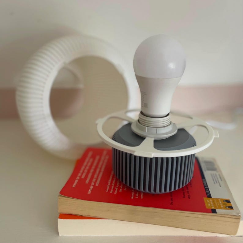 May include: A bright white LED light bulb is mounted on a grey and white fixture, resting on a stack of books with red and orange covers. A white, circular, ribbed object is in the background.