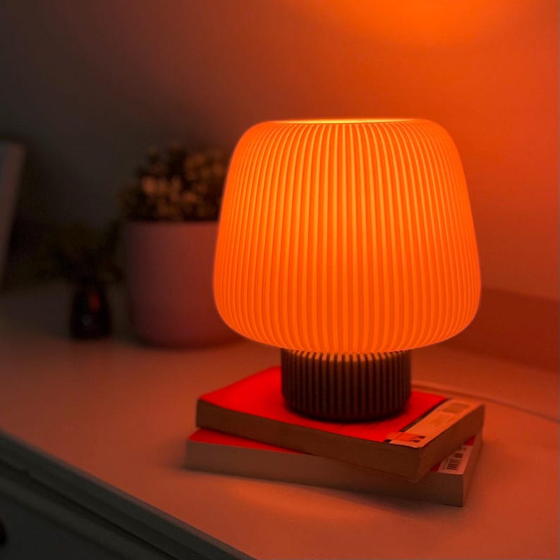 May include: An orange, ribbed table lamp with a dark brown base sits on two red books. The lamp emits a warm glow, casting an orange hue. A small potted plant is visible in the background. The lamp is a decorative item.