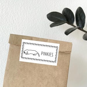 Könnte beinhalten: Eine braune Papiertüte mit einem weißen Etikett. Das Etikett zeigt eine schwarze Strichzeichnung eines Maulwurfs und das Wort "PINKIES". Das Etikett ist mit einem dekorativen Rand versehen. Im Hintergrund ist eine dunkelgrüne Pflanze zu sehen.