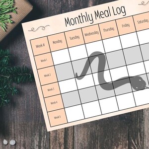 Puede incluir: Un planificador de registro de comidas mensual con una ilustración de serpiente. El planificador tiene un diseño de cuadrícula con los días de la semana y los números de semana. El fondo es una superficie de madera con una caja de regalo y vegetación.