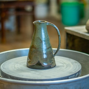 Puede incluir: Una jarra de cerámica hecha a mano con un asa curva. La jarra está esmaltada en tonos verdes y marrones, con una base acampanada. Se encuentra sobre un torno de alfarero, lo que sugiere que es una pieza de arte funcional. El fondo está borroso, centrándose en la jarra.