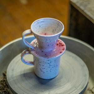 Pode incluir: Um conjunto de café para servir em cerâmica artesanal. O conjunto inclui uma caneca e um cone de servir, ambos em um esmalte branco e rosa salpicado. A caneca e o cone são empilhados. O conjunto está sobre uma superfície cinza.