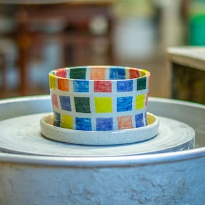 Puede incluir: Una colorida maceta de cerámica con un patrón de cuadrados rojos, azules, amarillos y verdes. La maceta está sobre un platillo blanco, centrado en un torno de alfarero. La escena tiene lugar en un taller.