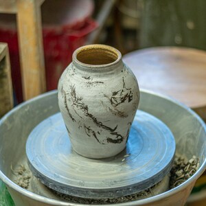 May include: A handmade ceramic vase with a textured, off-white surface and dark gray abstract patterns. The vase sits on a pottery wheel, surrounded by tools and other pottery equipment. The interior of the vase is a warm brown color.