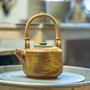 Może przedstawiać: Ręcznie wykonany ceramiczny czajnik z bambusowym uchwytem i małą, okrągłą pokrywką. Czajnik ma ciepły, ziemisty kolor z teksturowaną powierzchnią. Stoi na kole garncarskim.