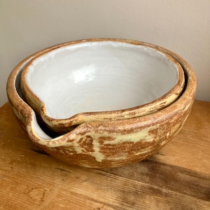 Puede incluir: Dos cuencos de cerámica para mezclar anidados. Los cuencos tienen un exterior marrón claro y un interior blanco. Los cuencos tienen un pico en un lado. Los cuencos están sobre una superficie de madera.