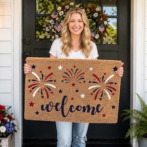 May include: A rectangular doormat with a brown background and a "welcome" message in navy blue script. The mat features red, white, and blue fireworks and star designs, perfect for a patriotic theme. The mat is displayed on a porch.