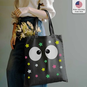 Puede incluir: Bolso tote negro con un dise&ntilde;o caprichoso de ojos blancos grandes y estrellas coloridas en amarillo, verde, rosa y blanco. El bolso tiene asas negras y est&aacute; hecho de una tela duradera. Ideal para llevar compras o art&iacute;culos personales.