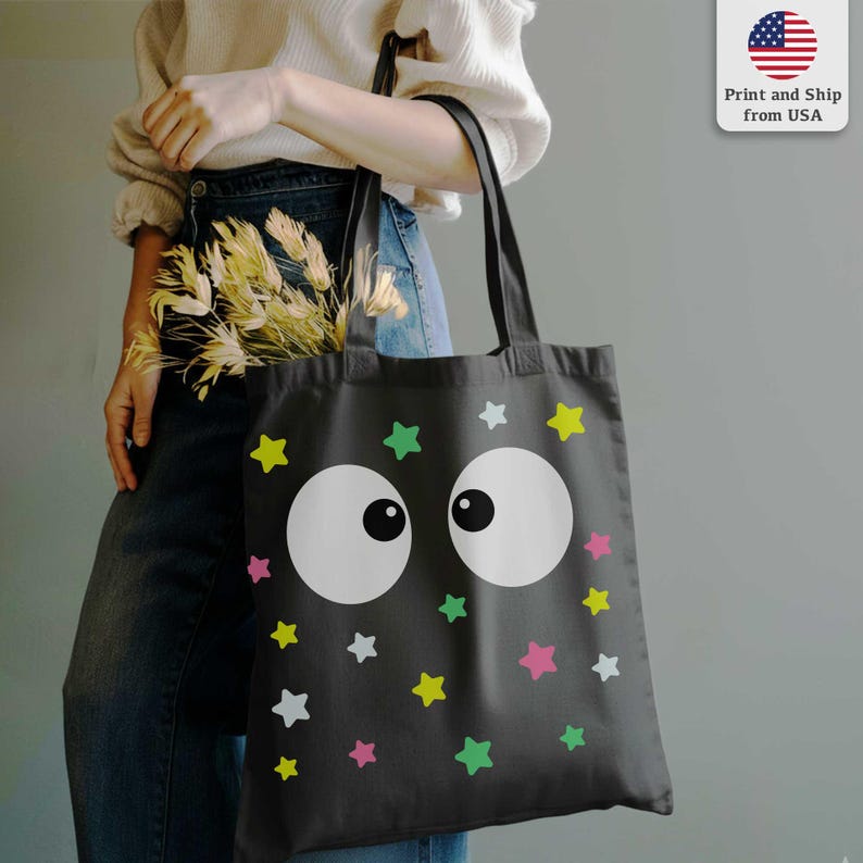 Puede incluir: Bolso tote negro con un dise&ntilde;o caprichoso de ojos blancos grandes y estrellas coloridas en amarillo, verde, rosa y blanco. El bolso tiene asas negras y est&aacute; hecho de una tela duradera. Ideal para llevar compras o art&iacute;culos personales.